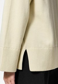 Licht beige gebreide trui met ribgebande mouwen en een zijsplit aan de zoom, die een gestructureerde stof en een ontspannen pasvormontwerp toont.