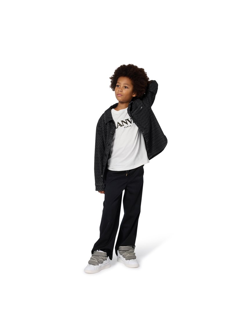 Schwarze gesteppte Jacke, weißes T-Shirt mit Logo, schwarze Hose und weiße Sneaker mit schwarzen Akzenten. Model posiert.