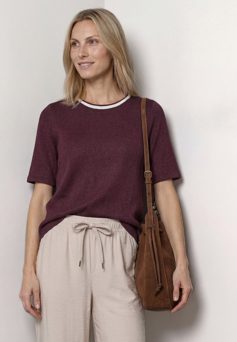Femme aux cheveux blonds mi-longs portant un haut bordeaux à manches courtes et un pantalon beige, avec un sac à bandoulière marron.