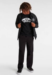 Zwarte Vans-jas over een zwart T-shirt met witte "Off the Wall"-grafiek, zwarte broek en zwarte schoenen. Gestructureerd materiaal, casual pasvorm.