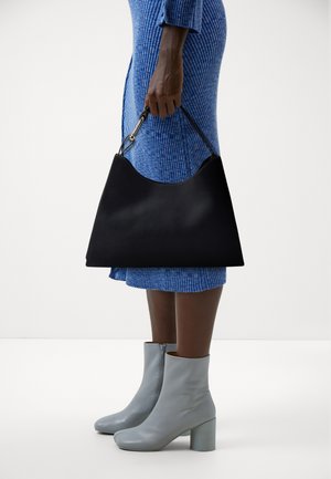 Sac en cuir noir avec un haut arrondi, tenu par une main. Porté avec une robe bleue à côtes et des bottines grises avec un talon épais.