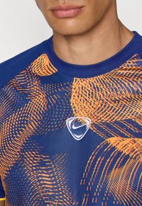Blue athletic shirt made of breathable fabric, featuring orange graphic patterns and a white Nike logo on the chest. Round neckline.