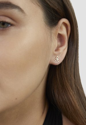 Nahaufnahme eines Damenohres mit einem kleinen goldenen Triquetra-Knoten-Stecker, der einen zentralen klaren Edelstein besitzt.