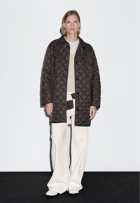 Hnědá prošívaná bunda s límcem a zapínáním na knoflíky, vrstvená přes světle zelený top, v kombinaci s širokými bílými kalhotami. Jednoduchý design, minimalistický styl.