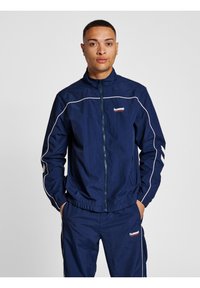 Navy blue windbreaker with white accents, zip closure, high collar, and elastic cuffs. Logo on the chest and chevron stripes on sleeves.