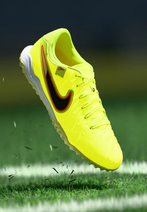 Jaskrawożółty but piłkarski Nike z czarnym i czerwonym logo, w powietrzu nad trawą i białymi liniami boiska.
