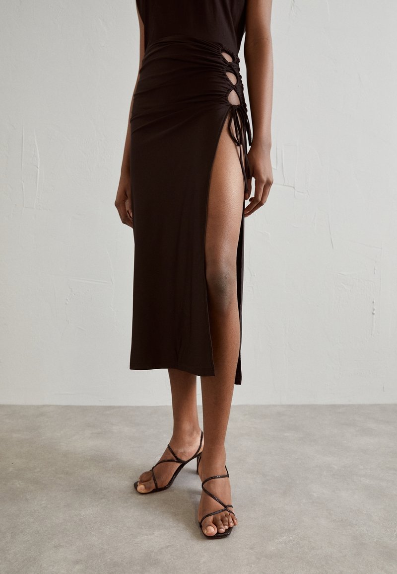 Brauner knielanger Rock mit gerafften Details und einem hohen Schlitz am Oberschenkel. Kombiniert mit Riemchensandalen mit offenen Zehen. Glattes, weiches Material.