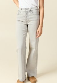 Mujer vestida con jeans de pierna ancha de tiro alto en color gris claro, camisa blanca metida por dentro y zapatos de ante marrón, de pie contra un fondo beige.