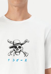 Hvid bomulds t-shirt med et sort kranie iført hat og korslagte knogler grafik, fremhævet af blå tekst nedenfor designet.