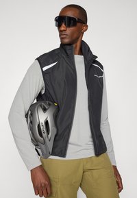 Schwarze isolierte Weste mit reflektierenden Akzenten, getragen über einem hellgrauen Langarmshirt. Grauer Helm unter dem Arm gehalten.