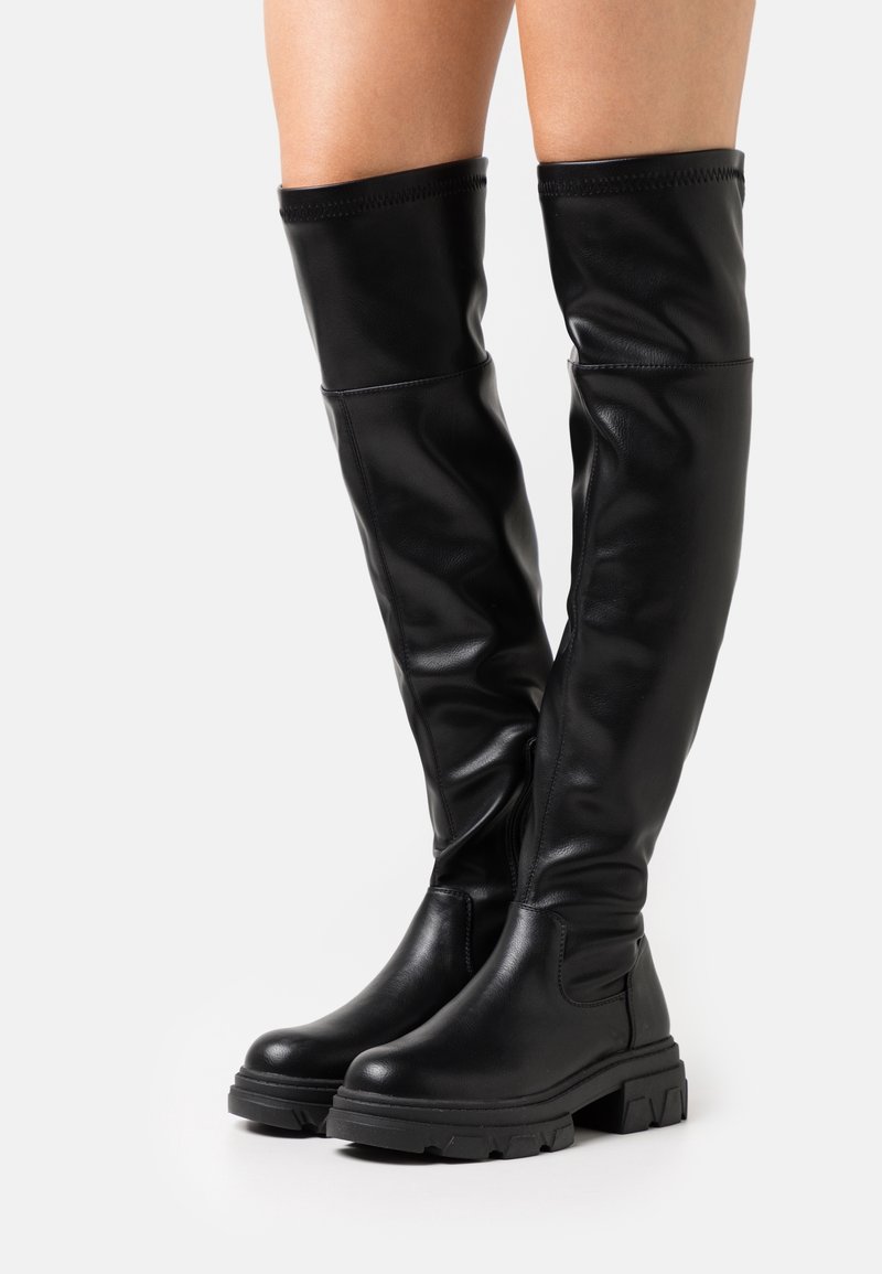 Bottes en cuir noires au-dessus du genou avec semelle épaisse et bout arrondi portées sur des jambes nues sur un fond blanc.