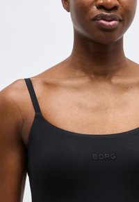 Κοντινό πλάνο ενός ατόμου που φοράει μαύρη αμάνικη μπλούζα με λεπτές τιράντες και το λογότυπο "BORG" στο κέντρο του στήθους.