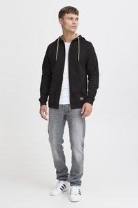 Nuori mies seisoo, yllään musta vetoketjullinen huppari, valkoinen t-paita, harmaat farkut ja valkoiset tennarit, joissa mustat raidat, katsoo oikealleen.