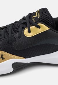 Gros plan d'une chaussure de sport noire et dorée avec tissu tissé, panneaux en cuir, lacets noirs et semelle blanche avec bande de roulement noire texturée.