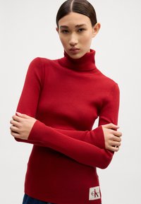 Rdeča rebrasta turtleneck pulover s dolgimi rokavi, z logotipom na spodnjem levem delu. Gladka tekstura in prilegajoč dizajn.