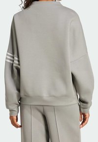 Felpa grigia chiara con vestibilità comoda, caratterizzata da tre strisce bianche sulla manica sinistra e un orlo a coste. Texture liscia.