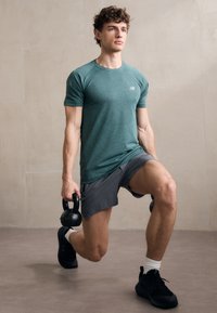 Joven con camiseta color teal y pantalones cortos grises realiza estocadas sosteniendo kettlebells negros en interiores.