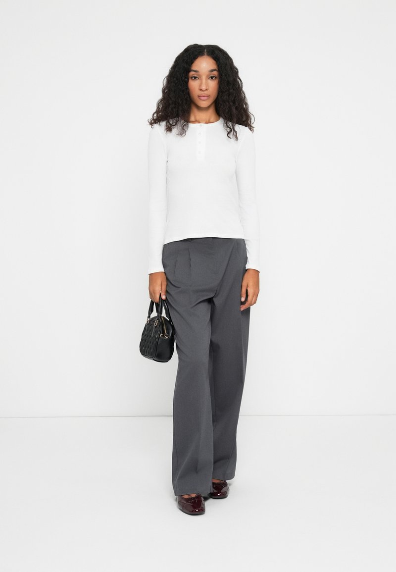 Haut blanc à manches longues avec un détail de boutons, associé à un pantalon gris taille haute et un petit sac à main noir. Chaussures vernies bordeaux.