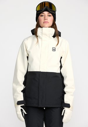 Veste de ski avec une partie supérieure blanche et une partie inférieure noire, dotée d'un col haut, d'une fermeture éclair sur le devant et de poignets réglables. Gants marron visibles.
