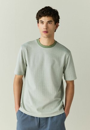 Junger Mann mit kurzen dunklen Haaren, der ein hellgrünes, strukturiertes Kurzarmhemd und blaue Hosen trägt, die Hände in den Taschen, schaut leicht nach rechts.