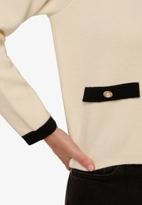 Waffelstrick-Pullover in Creme mit schwarzen Bündchen und einer Fronttasche, die mit einer goldenen und perlenförmigen Verzierung geschmückt ist.
