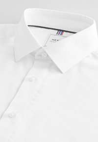 Chemise blanche à boutons avec col pointu et étiquette de marque visible à l'intérieur du col, conçue pour une tenue formelle ou professionnelle.
