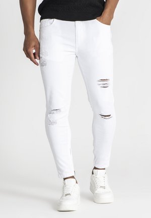 White ripped skinny jeans with distressed detailing, five pockets, and a tapered fit, paired with white sneakers.