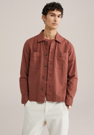 Rust-colored button-up shirt with two chest pockets, made of textured fabric. Paired with light-colored pants, features a casual collar.