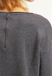 Szary dzianinowy sweter z żebrowanymi akcentami na ramionach oraz zapięciem na zamek z tyłu. Miękka tekstura i casualowy design.