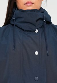 Woman wearing a navy blue jacket with a high collar and adjustable drawstrings, fastened with three visible round buttons.