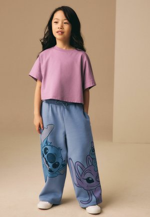 Młoda dziewczyna ubrana w luźny fioletowy t-shirt i szerokie niebieskie spodnie z postaciami z kreskówek, stojąca z jedną ręką w kieszeni na beżowym tle.