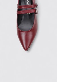 Zapatos Mary Jane de cuero burdeos con punta afilada, que presentan dos cierres de correa y hebillas en tono plateado. Textura suave y diseño elegante.