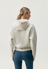 Felpa con cappuccio in beige chiaro, caratterizzata da un design attillato, polsini e cuciture a coste, abbinata a leggings blu scuro. Texture liscia.