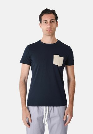 Uomo che indossa una maglietta blu navy con taschino beige sul petto e pantaloni grigio chiaro con coulisse bianche, in piedi davanti a uno sfondo semplice.