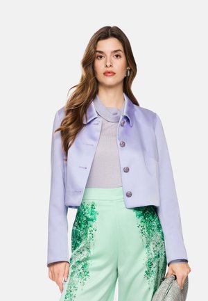 Woman with long wavy hair wearing a lavender cropped jacket, light gray top, and light green pants with dark green floral patterns, holding a textured bag.