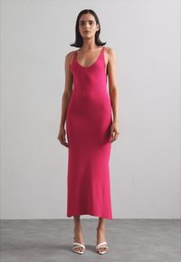 Abito aderente senza maniche, lungo fino al ginocchio, in tessuto a maglia fucsia vibrante, caratterizzato da una texture liscia e un design minimalista, abbinato a tacchi bianchi.