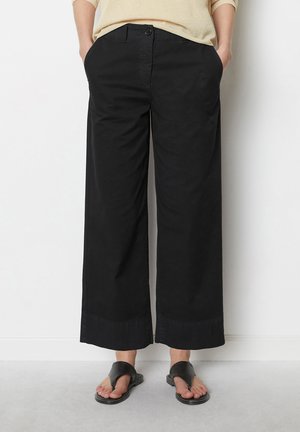 Person med løse sorte cropped bukser og sorte sandaler med åbne tæer, stående med hænderne i lommerne mod en hvid væg.