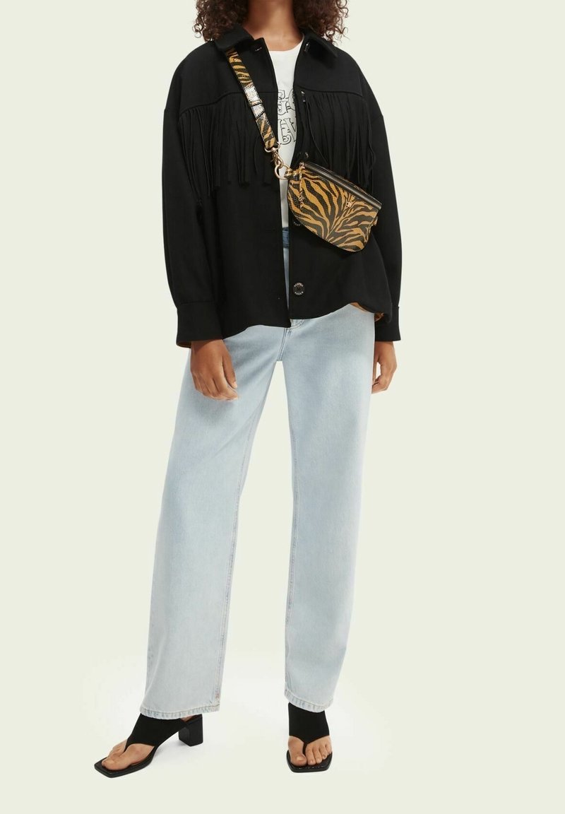 Veste-chemise noire à franges, t-shirt blanc avec motif, jean ample bleu clair et sandales noires. Accessoire : petit sac noir avec imprimé tigre orange.