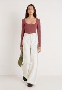 Pikkade varrukatega lavendlililla top laia kaelusega, kombineeritud laiade valgete pükstega. Käes väike oliivroheline käekott, jalas mustad kingad.