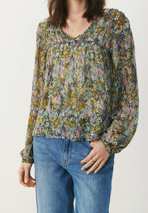 Kvinde iført en bluse med lange ærmer med blomstermønster i blå, pink og sennepsfarver samt blå jeans, hånd hvilende i lommen.