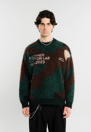 Jeune homme aux cheveux bouclés foncés portant un pull camouflage vert et marron avec un texte et un collier de perles, debout devant un fond uni.