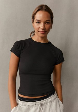 Jonge vrouw draagt een zwarte nauwsluitende top met korte mouwen en lichtgrijze joggingbroek met handen in de zakken tegen een effen grijze achtergrond.