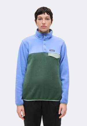 Person iført en blå og grøn fleece-pullover med høj knappet krave og en brystlomme, stående mod en ensartet lys baggrund.