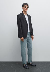 En ung man står mot en ljusgrå bakgrund, klädd i en svart blazer, vit skjorta, ljusblå jeans och solglasögon.