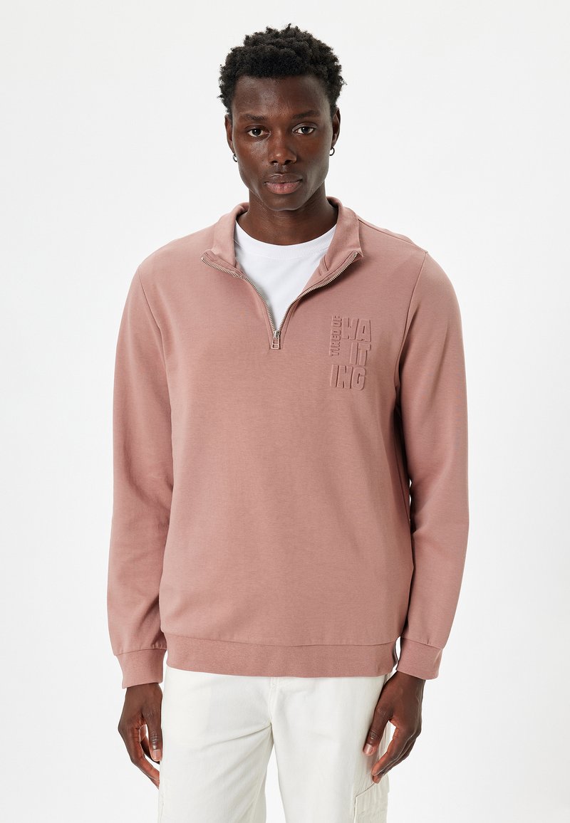 Sudadera de cremallera en rosa claro con un cuello alto, que presenta un diseño de logo texturizado en el pecho y mangas largas. Tejido suave y liso.