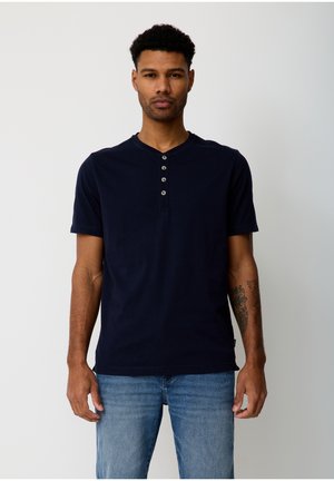 Mann mit kurzen lockigen Haaren, der ein marineblaues Henley-Shirt mit kurzen Ärmeln und hellblaue Jeans trägt und vor einem einfarbigen Hintergrund steht.