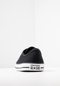 Musta kangaskengät pyöreillä sivuilla, valkoinen kumipohja ja musta-valkoiset raidat. Sisältää "Converse All Star" -logon kantapäässä.