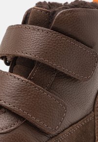Bota de cuero marrón con dos correas de Velcro, acentos de ante, acabado texturizado y forro suave, diseñada para comodidad y soporte.