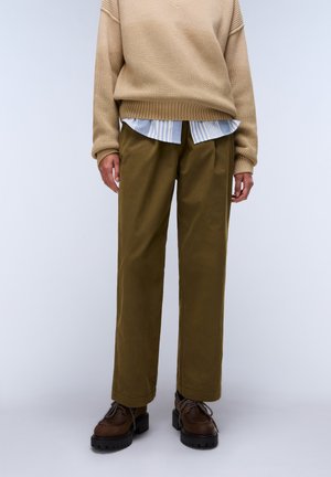 Person iført olivengrønne, lige bukser, brune chunky sko, beige strikket sweater og blåstribet skjorte synlig under.