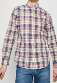 Man die een geruit overhemd met lange mouwen draagt in blauw, rood, groen en crème, gecombineerd met een blauwe spijkerbroek, staand met ontspannen armen.